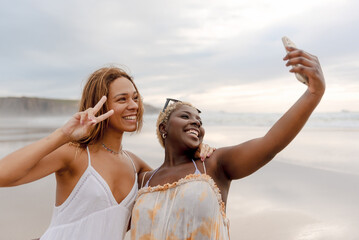 two friends having a good time on the beach taking selfie pictures with their smart phone during a...