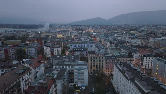 Aerial panoramic view of city at dusk. Forwards fly above residential multistorey buildings in urban borough. Mountains and famous Jet d Eau fountain in background. Geneva, Switzerland
