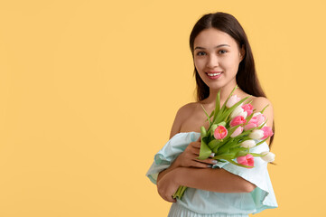 Happy young Asian woman with bouquet of beautiful tulips on yellow background. International...