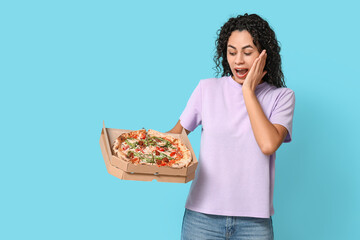 Beautiful young shocked African-American woman with delicious pizza on blue background