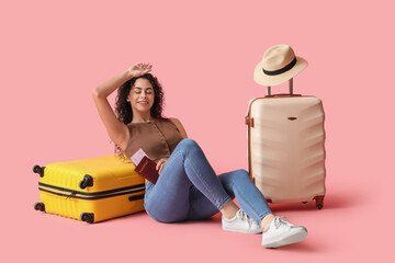 Young African-American female tourist with suitcases and passport sitting on pink background
