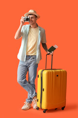 Young male tourist with suitcase, photo camera and passport on orange background