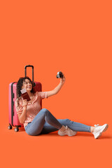 Fototapeta premium Young African-American female tourist with suitcase and passport taking selfie on orange background