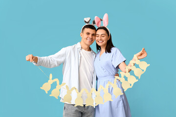 Happy young couple with Easter bunny ears and garland on blue background
