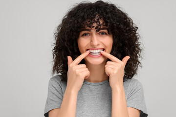 Young woman applying whitening strip on her teeth against light grey background