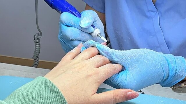 Specialist Uses a Milling Machine to Remove Gel Polish, the Master does a Hardware Manicure in Protective Gloves under the Light of a Lamp, Particles of the Old Coating Scatter into Dust under Light