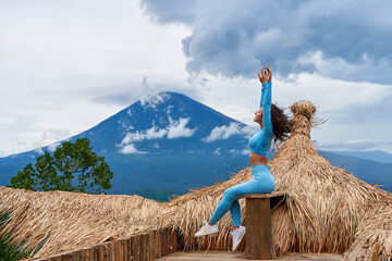 A young woman poses on a large straw photo zone as a bird on a viewing platform and enjoys the view of the sacred Mount Agung volcano hidden by clouds on a rainy day on the island of Bali.