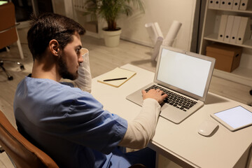 Male nurse with laptop working evening shift at table in clinic