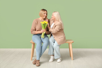 Adult woman with her mother and bouquet of tulips sitting on bench near green wall. International...
