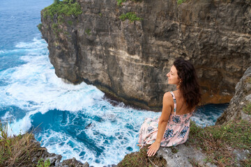 A beautiful woman in a pink dress sits on a cliff above the ocean on the island of Nusa Penida. Devil's Billabong an incredibly wonderful lagoon with splashes from the waves.