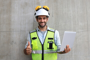 A smart engineer is standing in front of a cement wall, holding a laptop.