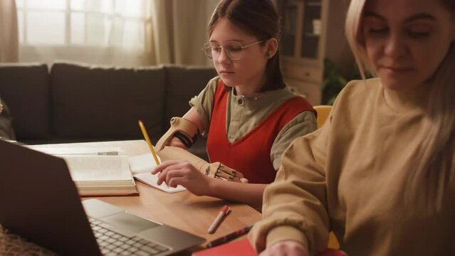 Side footage of Caucasian homeschooler girl with prothesis doing schoolwork and using laptop computer under mothers assistance