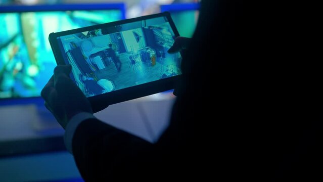 Man security guard officer working on tablet in monitoring center, zooming watching video. Computers and big digital screen show cctv cameras footage.