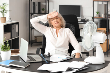 Mature businesswoman suffering from hot flashes at table in office. Menopause concept