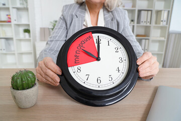 Mature businesswoman holding clock with word MENOPAUSE in office, closeup