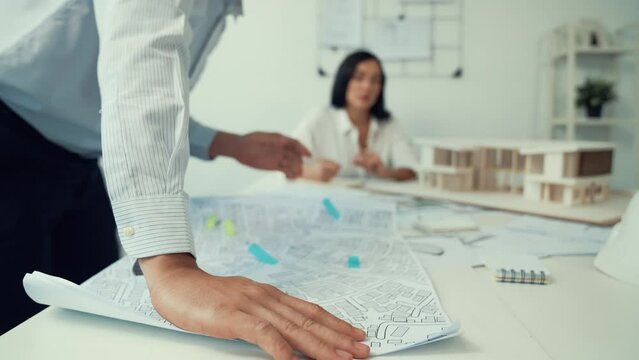 Skilled caucasian interior designer working with house model while discussing with asian architect engineer about house plan on meeting table with blueprint and house model scatter around. Immaculate.