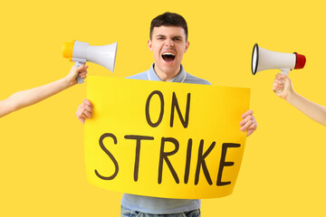 Protesting young man holding placard with text ON STRIKE and megaphones against yellow background