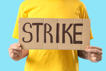 Protesting young man holding placard with word STRIKE on blue background, closeup