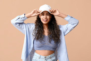 Stylish African-American woman in shirt, jeans and cap on beige background