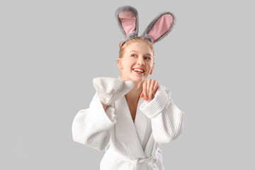 Happy young woman in bathrobe with bunny ears and massage glove on grey background