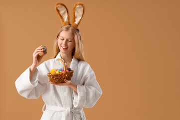 Young woman in bathrobe holding basket with Easter eggs and bath sponge on color background