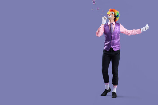 Portrait of clown blowing soap bubbles on lilac background. April Fool's day celebration