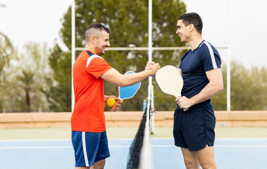 Two grown men are playing a game of pickleball on an outdoor court.The boys high-five each other to greet each other happily.Pickle ball concept. Men Playing Pickleball - obrazy, fototapety, plakaty