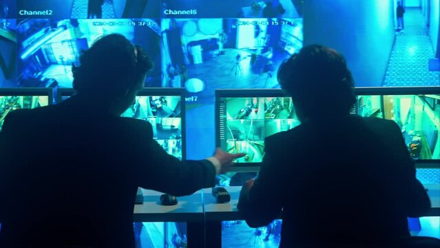 Male security guard officers working on pc in monitoring center, discussing and speaking by radio. Computers and big digital screen show cctv cameras footage.