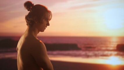 Profile of a young man with bare torso admiring a sunset, his face illuminated by the warm golden...