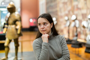 Portrait of interested adult female tourist visiting exhibition of medieval armor in armory of historical museum ..