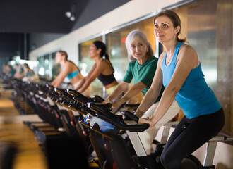 Positive mature female riding exercise bike during cycling class in modern gym