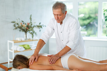 Experienced male massage therapist giving general back massage to female patient lying at massage table