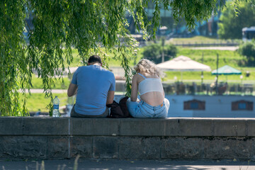 A man and a woman sit side by side under the shade of a tree on a sunny day, engrossed in their...