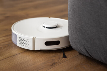 A white robot vacuum cleaner cleans a dark oak wood laminate on the floor. collects dust near the...