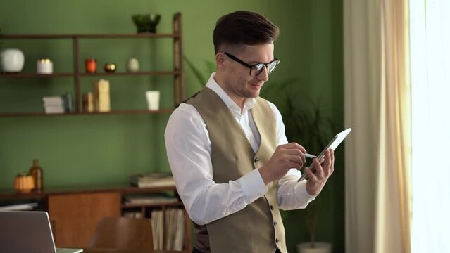 A male manager is a creative individual with glasses and a shirt, who works from home using a tablet at his workplace.
