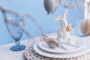 Table place setting with bunny figure and eggs, empty glasses and festive easter decor on pastel...