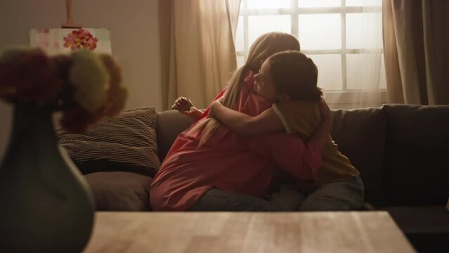 Side slowmo of mother supporting and hugging her little daughter with prosthetic arm while sitting on sofa in cozy living room