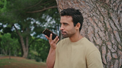 Italian man recording voice message on smartphone leaning at tree trunk close up