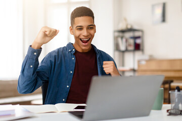 Joyful black teen guy celebrating success in front of laptop