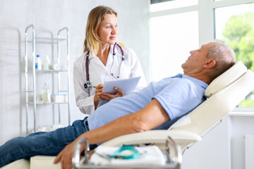 Fototapeta premium Positive female doctor conducting questionnaire of elderly male patient lying on couch in medical office, asking series of questions and noting down responses in document folder