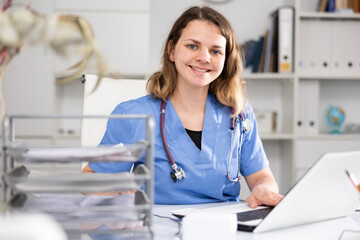 Portrait of female doctor in surgical scrubs sitting at working table in her office.