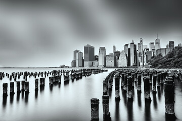 Black and white photo of the iconic manhattan skyline over calm waters, with weathered pier remains in the foreground