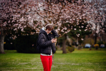 Engrossed in her smartphone and music, a young woman stands under the spring bloom of cherry...