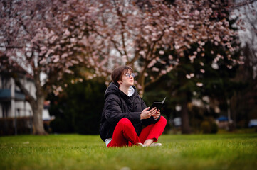 A focused young woman sits cross-legged on the grass in a park, deeply engaged with her tablet with...