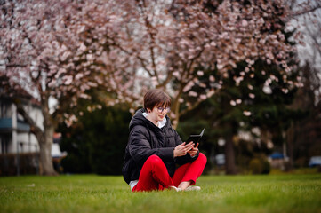 A focused young woman sits cross-legged on the grass in a park, deeply engaged with her tablet with a beautiful cherry blossom tree flowering in the background