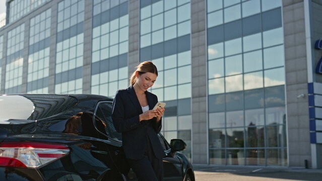 Happy business woman texting on cellphone leaning at luxury car outdoors zoom in