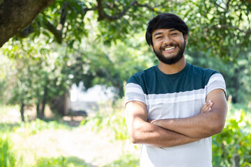 Naklejka na ściany i meble Portrait of young happy smiling Indian asian bearded man model standing at outdoor park looking confident with crossed arm