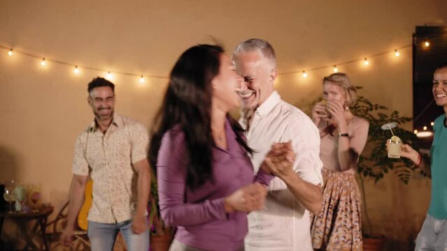 Multiracial adult attractive couple having fun dancing hand in hand with friends cheering at private backyard party home. Happy and joyful mature people celebrating outdoor gathering on summer evening