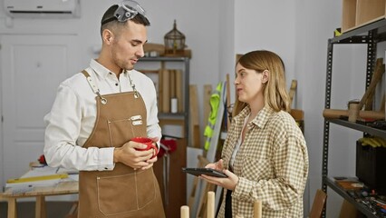 Man and woman discuss woodworking projects in a well-equipped indoor workshop.