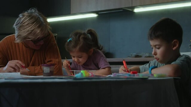 Cheerful elderly woman and two kids sitting at table and drawing with multicolored markers on paper. Good time together